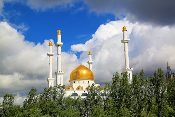 Mosque with golden dome and white minarets rising above trees against dramatic cloudy sky.