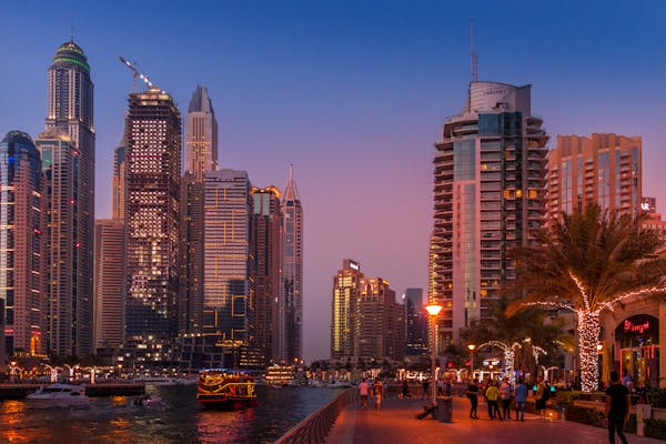 Dubai Marina skyline at dusk with illuminated skyscrapers, palm trees with lights, and boats on the water as pedestrians stroll along the waterfront promenade.