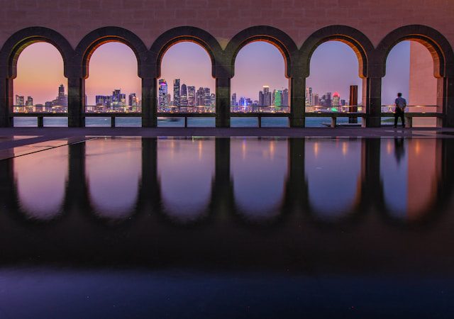 Doha skyline at sunset viewed through arched colonnade with reflections in water.